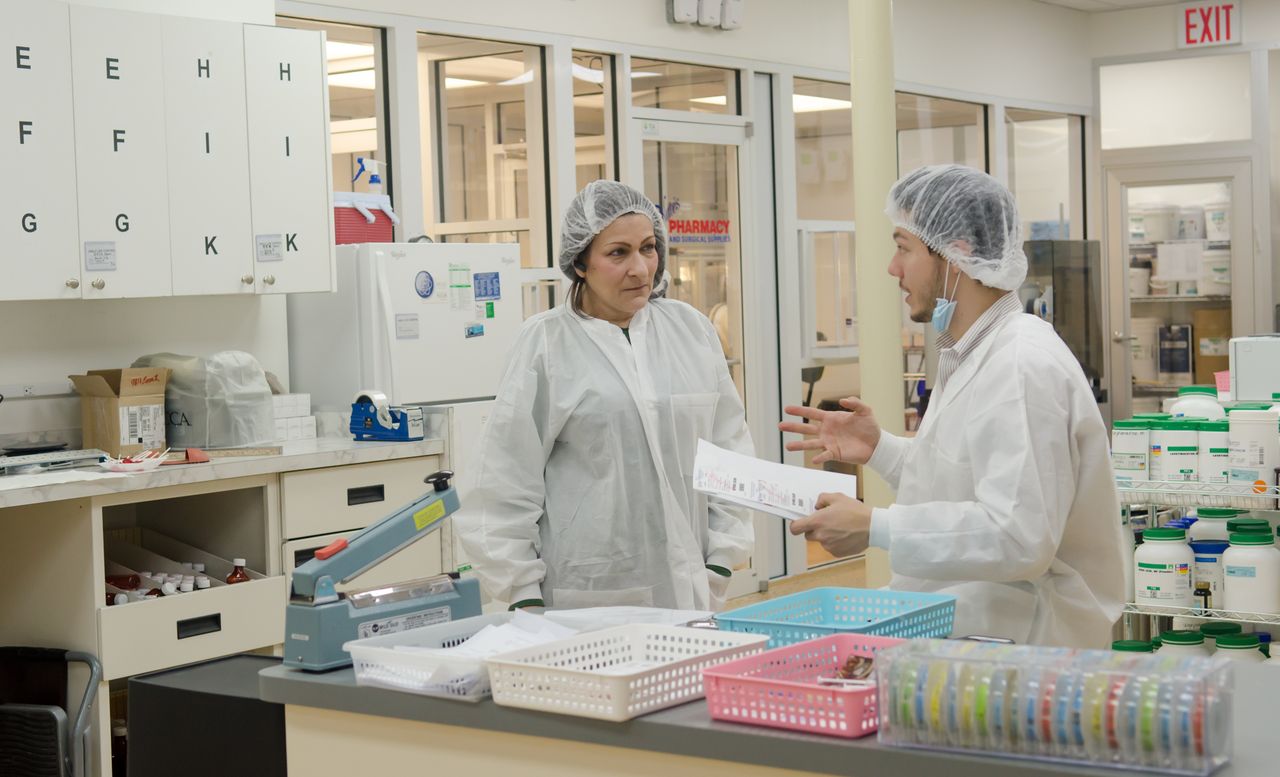 About Concentrated laboratory workers wearing white robes and hats standing with papers in contemporary chemical room and discussing results of scientific research